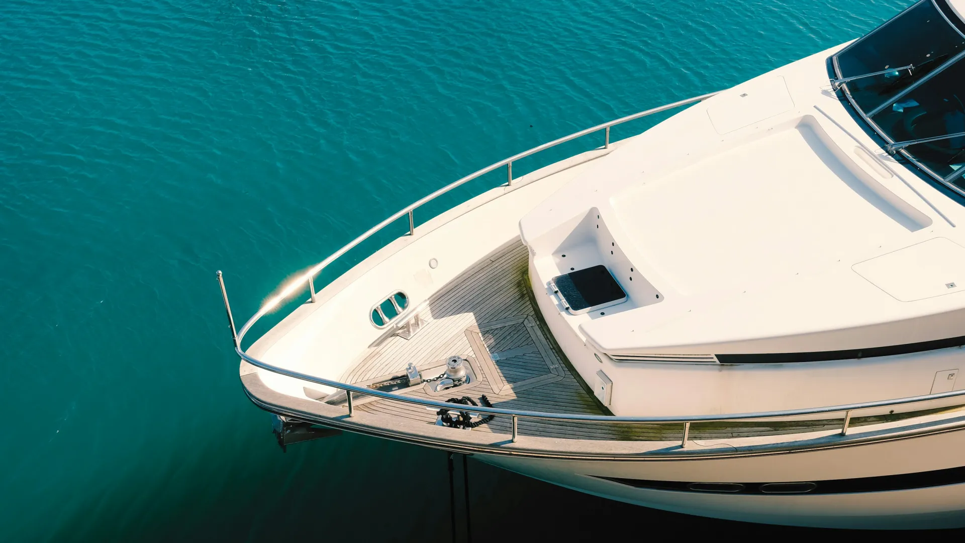 Close-up of a white yacht bow floating on clear turquoise water with metal railings and wooden deck visible