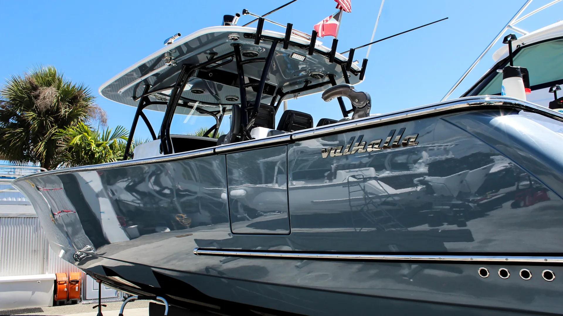 Glossy gray Valhalla boat with flags docked under clear blue sky near palm trees.