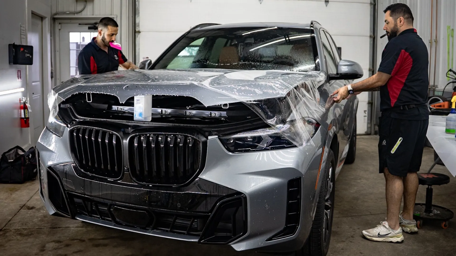 Two men applying protective film to the hood of a silver BMW SUV inside a garage.