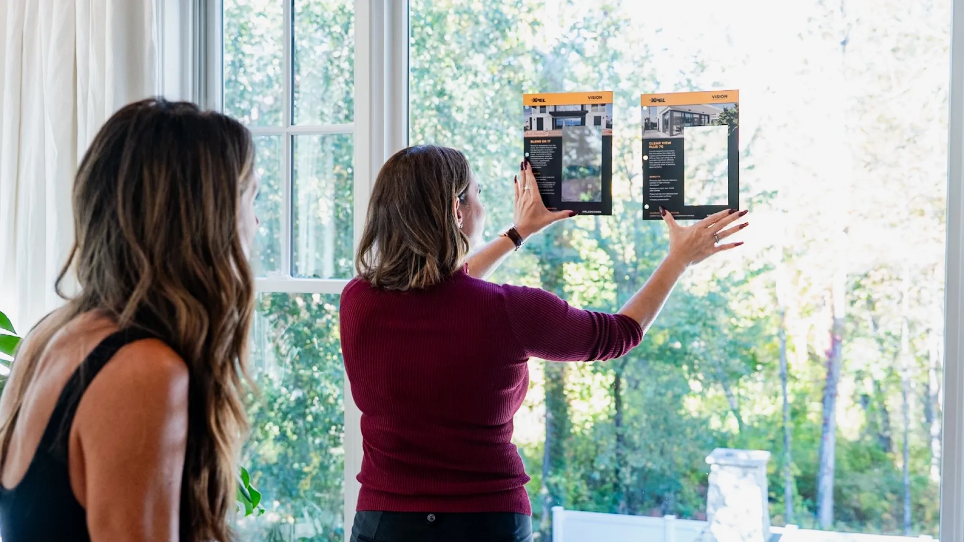 Two women comparing window sample clips against a large bright window with a green outdoor view.