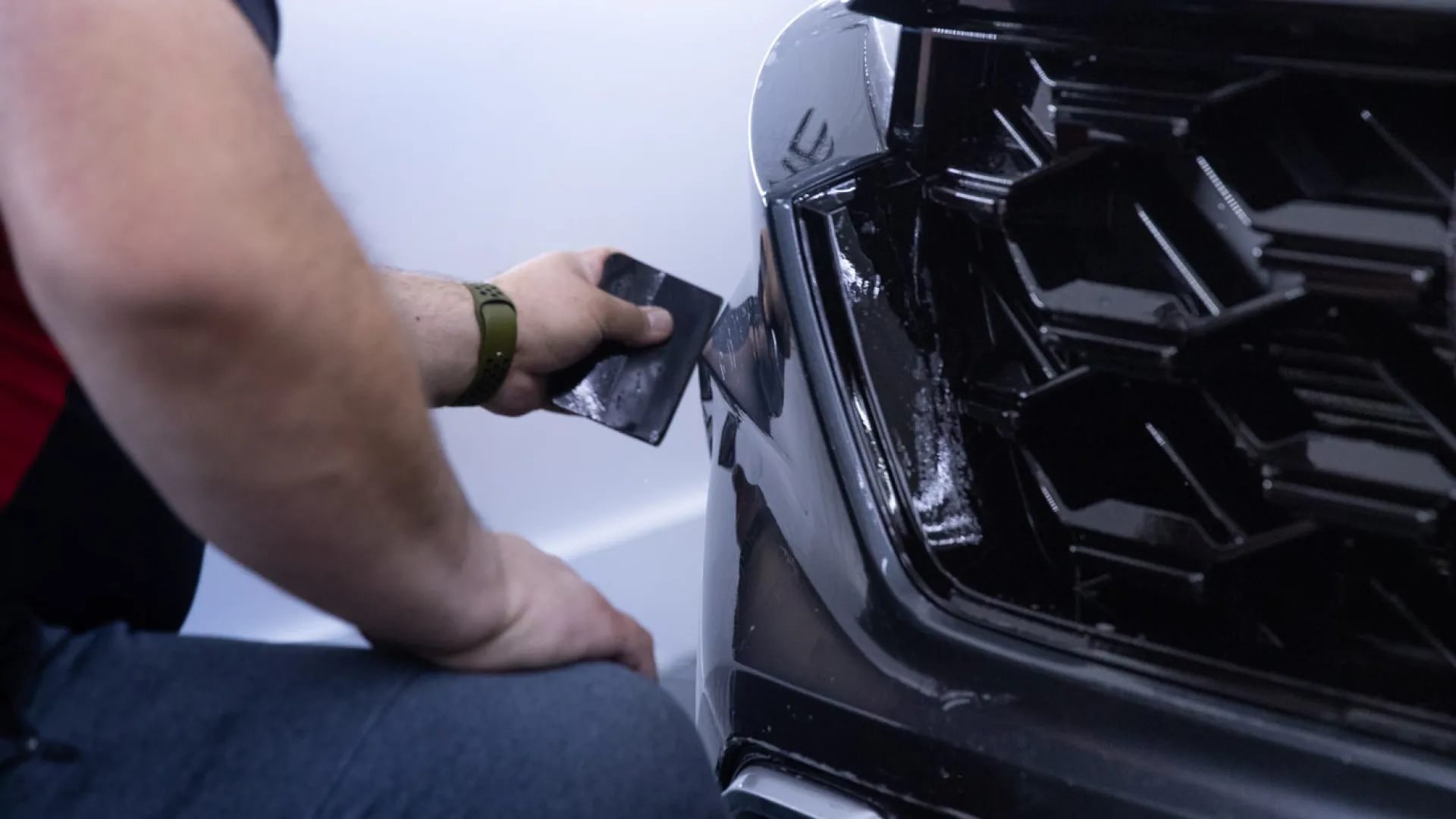 Technician applying vinyl wrap or protective film to the front bumper of a black car in a workshop.