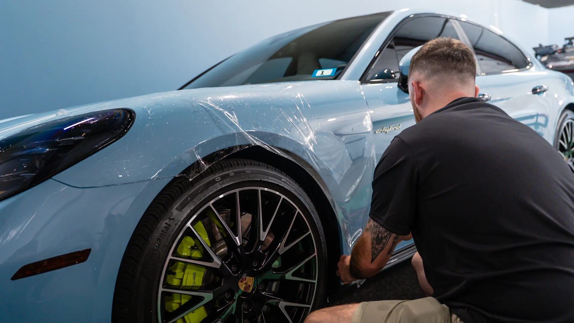Man applying protective film on light blue Porsche car's front left fender and wheel area indoors.