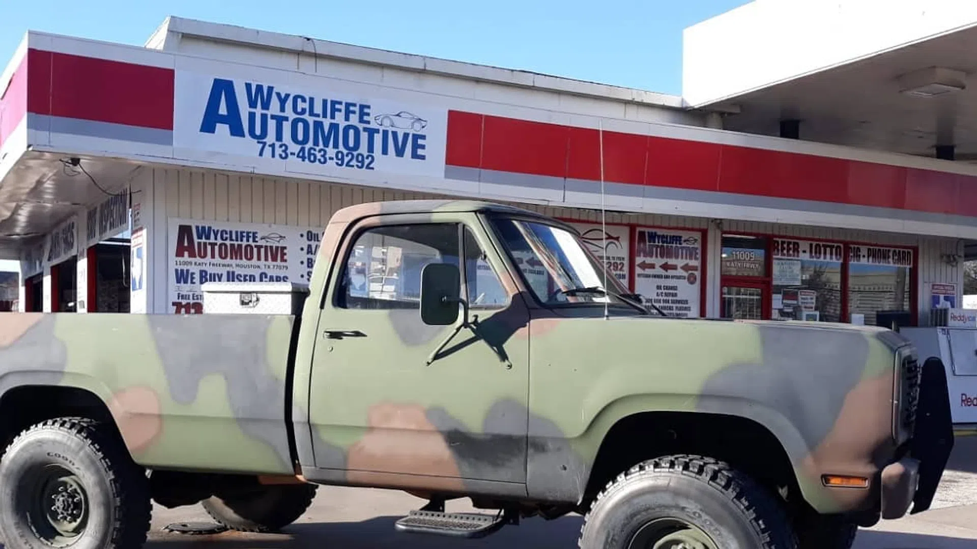 Camouflage-painted pickup truck parked outside Wycliffe Automotive with clear signage and blue sky.