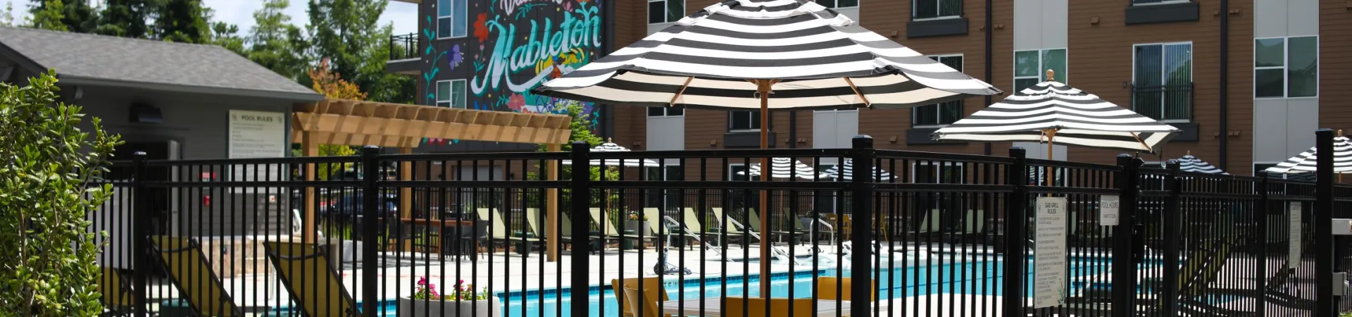 Apartment pool area with striped umbrellas, lounge chairs, and a 'Welcome to Mableton' mural on building.