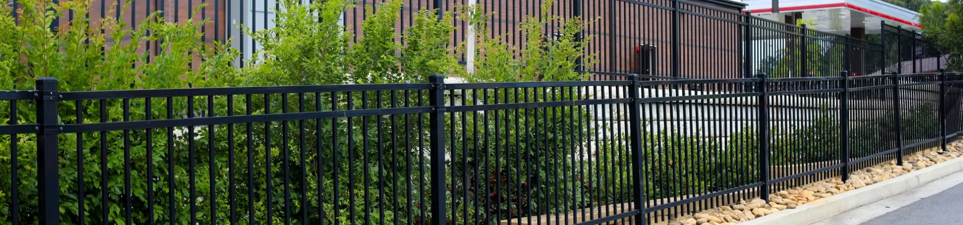 Black metal fence with green shrubs and rocks lining a sidewalk next to a brick building under a clear sky