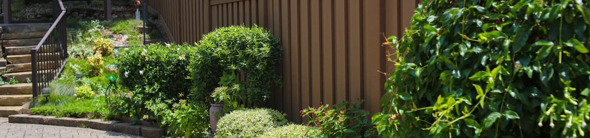 Brown wooden fence bordered by lush green shrubs and a paved patio walkway on a sunny day