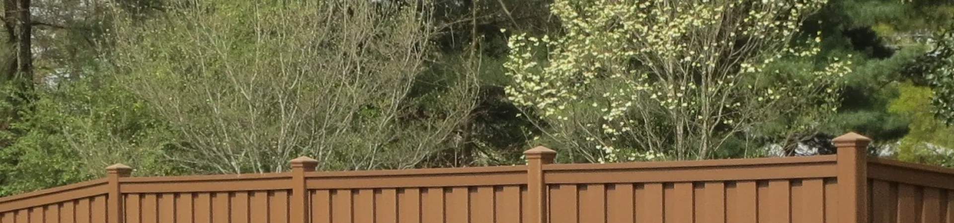 A brown vinyl fence surrounding a pool area, bordered by lush greenery and trees in a sunny outdoor setting.