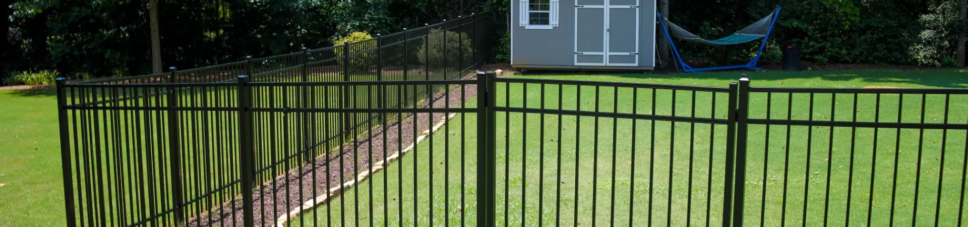 Black metal fence encloses green backyard with small shed and hammock under trees on sunny day