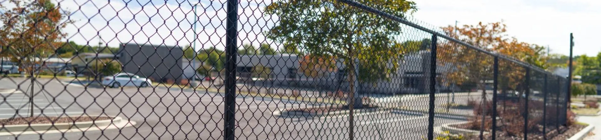 Chain-link fence along a smooth pavement near a parking lot with trees and clear blue sky.