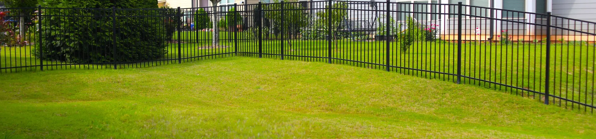 Green backyard with trimmed grass surrounded by black metal fence and houses in the background on a sunny day