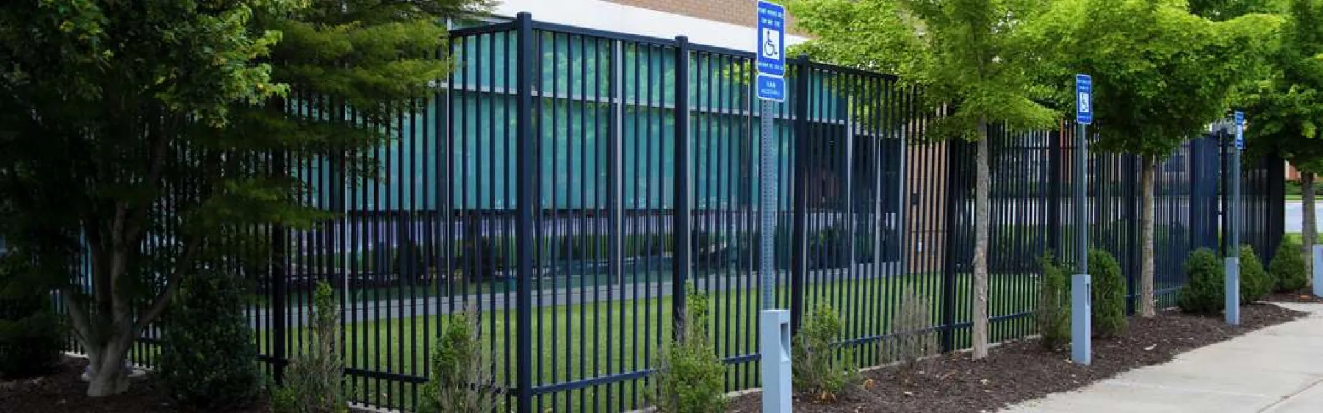 Black metal fence enclosing landscaped area with small trees and bushes along a sidewalk and handicapped parking signs.