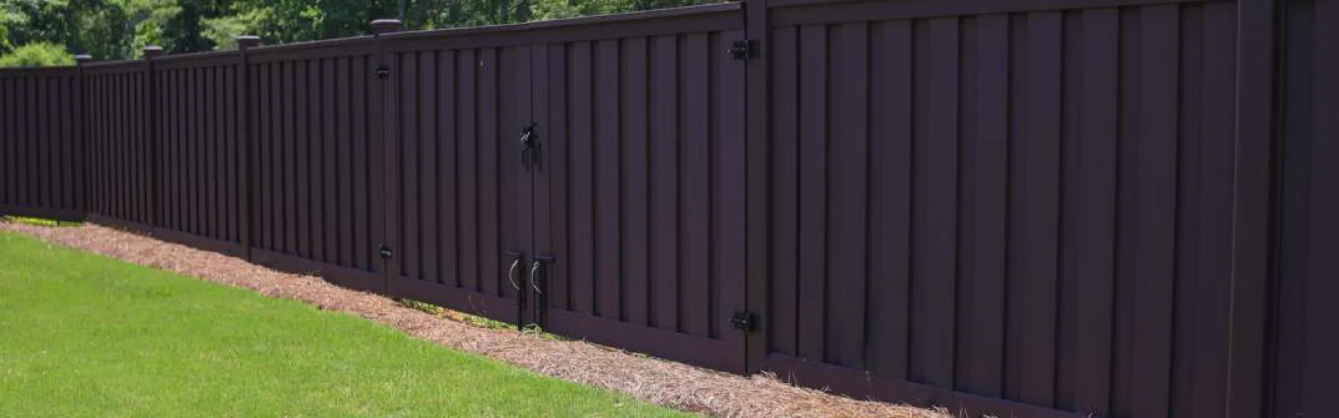 Long dark brown wooden fence with gate enclosing a grassy yard next to green trees in sunlight