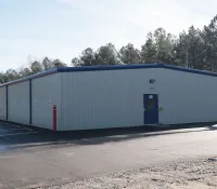 Single-story industrial metal building with blue trim beside a paved parking lot under clear sky near trees.