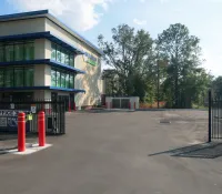 Modern storage facility with gated entrance, office building, and surrounding trees under blue sky.