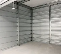 Empty storage unit with metal walls and a concrete floor, showing a partially open roll-up door and pull cord.