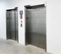 Two closed stainless steel elevator doors in a white indoor hallway with safety signs and buttons.