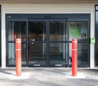 Automatic sliding glass doors with red bollards and a keypad access system outside a storage facility.