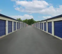 Outdoor self storage units with blue roll-up doors lining a paved driveway under a partly cloudy sky.