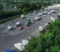 Large parking lot with various RVs, trailers, and vehicles surrounded by trees and nearby buildings on a clear day