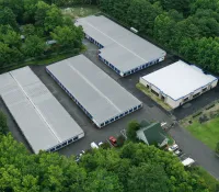 Aerial view of a storage facility with multiple large buildings surrounded by dense green trees and parked vehicles.