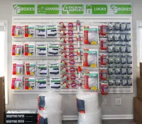 Wall display of moving and packing supplies including tape, covers, locks, and various boxes in a storage room.