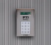 Digital security keypad mounted on a grey corrugated wall displaying date and time on screen.