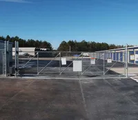 Chain link security gate closed at the entrance of a large outdoor storage facility with storage units and clear blue sky.