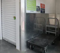 Storage unit hallway with closed white roll-up door, caution signs, and two black flatbed carts on concrete floor.