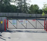 Closed black metal gate with signs and red bollards on paved entrance with wooden fence and trees in background