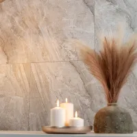 Three lit white candles on a tray beside a rustic vase with dried pampas grass against a stone wall.