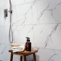Modern bathroom with marble wall tiles, wooden stool, folded towels, brown pump bottles, and handheld shower.
