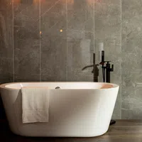 Modern white freestanding bathtub with towel against gray marble wall and wooden floor under warm light.