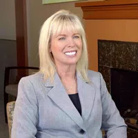 Smiling blonde woman in a gray blazer sitting in a cozy room near a brown fireplace with stacked logs.