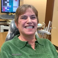 Smiling woman in green sweater sitting in a dental office chair with dental tools and computer screen in the background