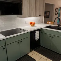 Modern kitchen with green lower cabinets, white countertops, black sink, stovetop, and a basket of oranges.