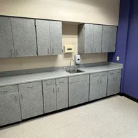 Modern kitchenette with grey cabinets, single sink, soap dispenser, and purple accent wall by a window.