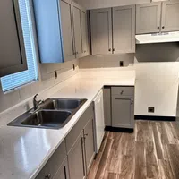 Modern kitchen with gray cabinets, white countertops, double stainless steel sink, and wood-look flooring.
