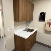 Small medical room sink with wood cabinets, white countertop, hand sanitizer dispenser, and medical waste bin.