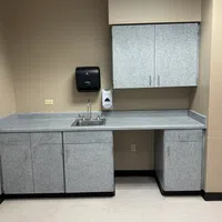 Empty utility room with gray cabinets, stainless steel sink, soap dispenser, and paper towel holder on beige walls.