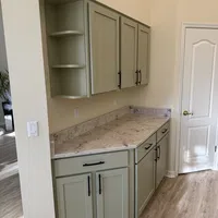 Kitchen corner with sage green cabinets, marble countertop, black handles, light wood flooring, and white door.