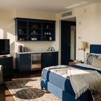 Cozy hotel room with navy blue bed, patterned bedding, kitchenette, TV, armchairs, and natural light from window.