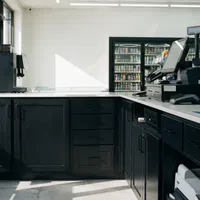 Modern convenience store counter with black cabinets, coffee machine, snack racks, and refrigerated beverages in background