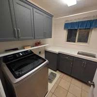 Laundry room with stainless steel washer, gray cabinets, white countertop, beige tiled floor, and window with blue valance