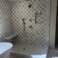 Modern shower with beige checkered tiles, gold fixtures, marble bench and a white freestanding bathtub.