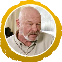 Bald man with a white beard wearing a beige shirt and chain necklace in a softly lit indoor setting.
