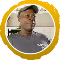 Smiling man wearing a dark cap and t-shirt with the name James, sitting indoors with kitchen in background.