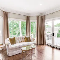 Bright living room with white sofa, beige curtains, glass coffee table, hardwood floor, and large windows with garden view.