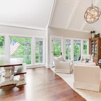 Bright living room with high white ceiling, beige armchairs, wooden flooring, fireplace, and large windows with garden view