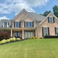 Two-story brick house with black window shutters, landscaped front yard, covered porch, and sloped green lawn.