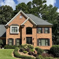 Two-story brick house with gray roof, black shutters, and landscaped garden under a blue sky with clouds.